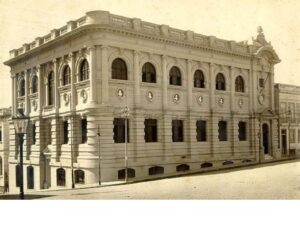 A instituição foi fundada em 14 de abril de 1871, durante o reinado de D. Pedro II, mas no seu início, a Biblioteca ocupava uma sala no Atheneu Rio-Grandense, na rua Duque de Caxias, onde funciona hoje a escola Bom Jesus Sevigné.
Em 1915, já autônoma, a Biblioteca foi transferida para a sede atual na rua Riachuelo, esquina General Câmara — conhecidas como rua do Cotovelo e Rua do Ouvidor. 
O prédio foi projetado pelos engenheiros das Obras Públicas do Estado Affonso Hebert e Teófilo Borges de Barros.
Tanto no exterior como em seu interior, a construção apresenta influência da doutrina positivista, utilizando vários estilos em sua representação. A fachada apresenta o estilo neoclássico, contornada com bustos do calendário positivista vindos da França, tais como Júlio César, Gutemberg e Descartes. Nas outras salas e salões, como o Salão Egípcio, Sala Borges de Medeiros e Salão Mourisco, diversificam-se os estilos, entre eles o rococó, egípcio, gótico e florentino. 
A maioria dos trabalhos foi executada por Fernando Schlater e S. Incerpi (pintores) e Alfred Adlof, Eduardo de Sá e Giuseppe Gaudenzi (escultores). O mobiliário, aberturas e objetos de adorno foram desenhados pelos mesmos artistas e fornecidos pelas firmas Jamardo Irmãos e João Friederichs S.A.
Victor Silva, diretor da BPE na época, orientou o projeto e a decoração do prédio. Ele teria se inspirado na Igreja de Sainte Geneviéve, em Paris, que havia sido transformada em biblioteca.
O prédio da BPE foi inaugurado como parte das comemorações do Centenário da Independência, a 7 de setembro de 1922, e é por isso que os vitrais das portas e janelas são na cor verde.
Projetada para ser um espaço nobre de leitura, então privilégio de elites, o espaço foi projetado para refletir o progresso, a força do Estado e a influência da doutrina Positivista, que detinha inúmeros seguidores no Rio Grande do Sul, com Júlio de Castilhos (1860 – 1903), à frente.
O próprio governador Borges de Medeiros (1863 – 1961) utilizava a Biblioteca para despachar e fazer suas leituras diárias de jornais na sala que hoje tem seu nome.
Era comum a elite cultural e política gaúcha usar a BPE como ponto de encontro para discussões políticas e intelectuais, sediando apresentações de música de câmara, conferências e saraus poéticos. Porto Alegre almejava conquistar “o espírito da época”, como importante polo intelectual e político elite cultural, nos moldes da Biblioteca Nacional, no Rio de Janeiro. 
Em 1986 o prédio foi tombado pelo Instituto do Patrimônio Histórico e Artístico do Estado (IPHAE), e em 2000 pelo Instituto do Patrimônio Histórico Nacional (IPHAN)  e é considerado um dos maiores tesouros arquitetônicos dos gaúchos.
Venha visitar a Biblioteca Pública do Estado e fazer parte dessa história! O funcionamento é de segunda a sexta, das 10h às 18h, e aos sábados, das 10h às 17h.
[/caption]
									</div>

																			<div class=